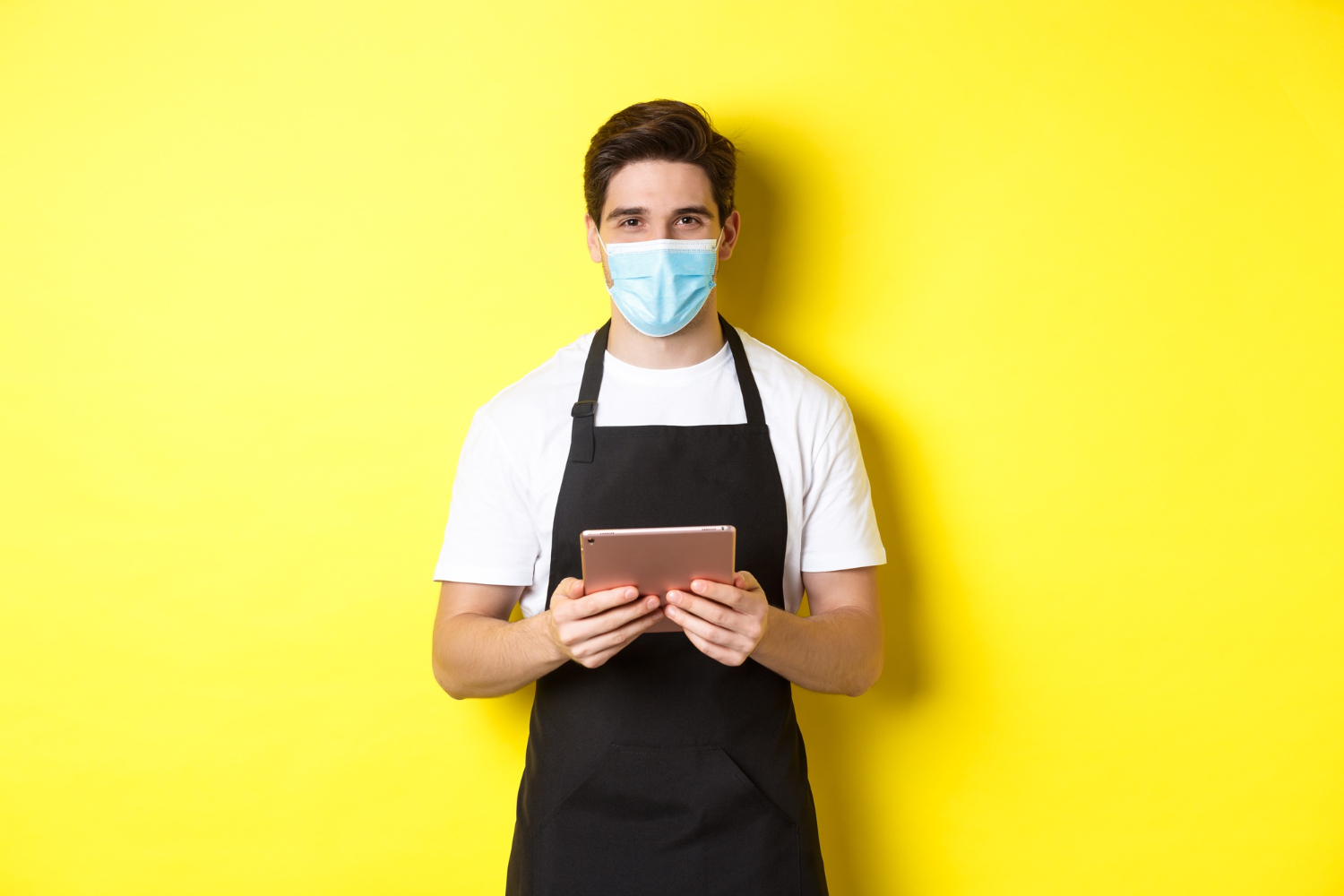 concept of covid small business and pandemic waiter in black apron and medical mask taking order holding digital tablet standing over yellow background